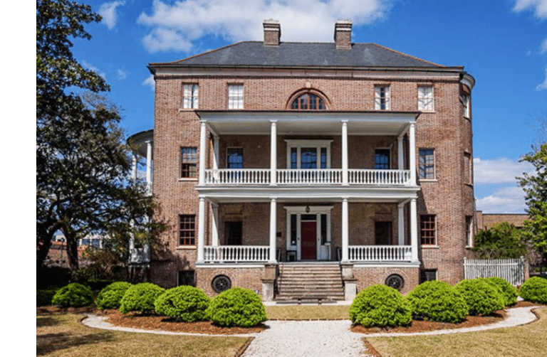 This large brick house, reminiscent of a classic Charleston estate, features two-story white porches with stately columns and a central staircase. It's surrounded by manicured bushes, a graceful tree, and set against a partly cloudy sky, capturing the essence of Southern charm.