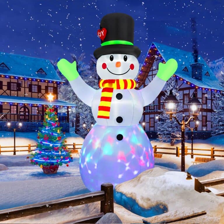 The inflatable snowman with a top hat and scarf stands proudly as part of the enchanting Christmas outdoor decorations in a snowy village scene, surrounded by lit houses and a shimmering Christmas tree at night.