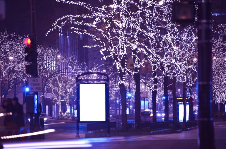 City street at night with trees adorned in bright white lights. Illuminated sign and blurred car lights visible, creating a festive ambiance.