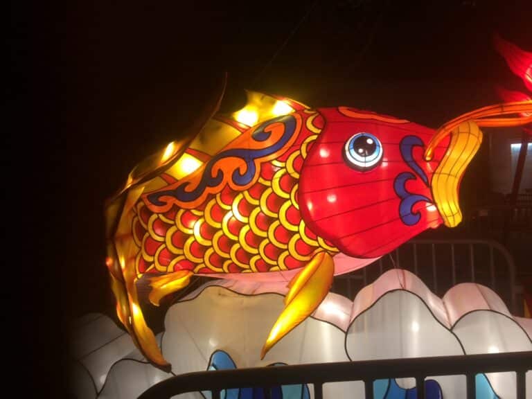 Illuminated lantern in the shape of a colorful koi fish with intricate patterns and bright colors against a dark background.