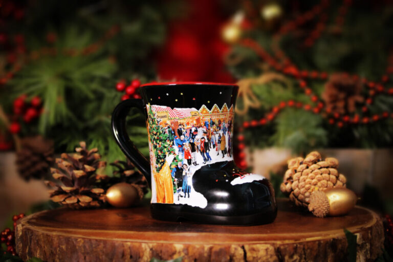 A boot-shaped mug with a festive holiday market scene sits on a wooden surface, surrounded by pinecones and greenery.