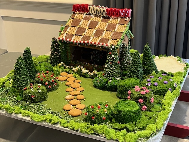 A gingerbread house with a decorated roof sits in a landscape of green frosting, trees, and small round cookies forming a path. Vibrant flowers and bushes surround the scene.