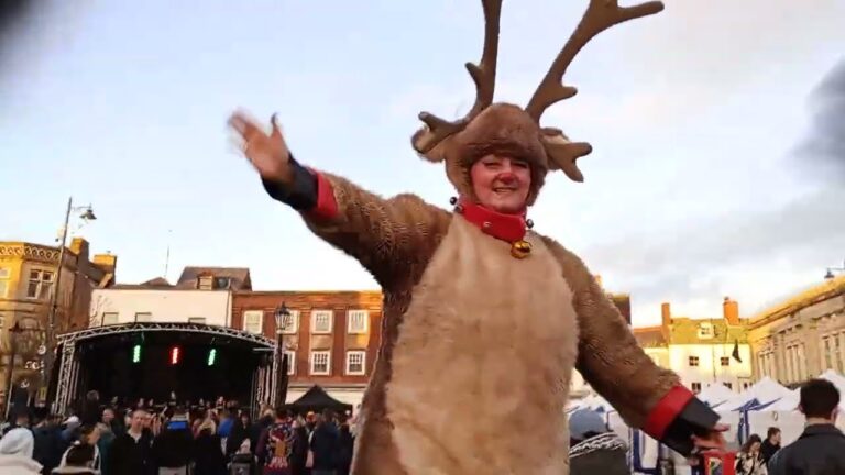 A person in a reindeer costume waves to a crowd at an outdoor event, with a stage and buildings visible in the background.