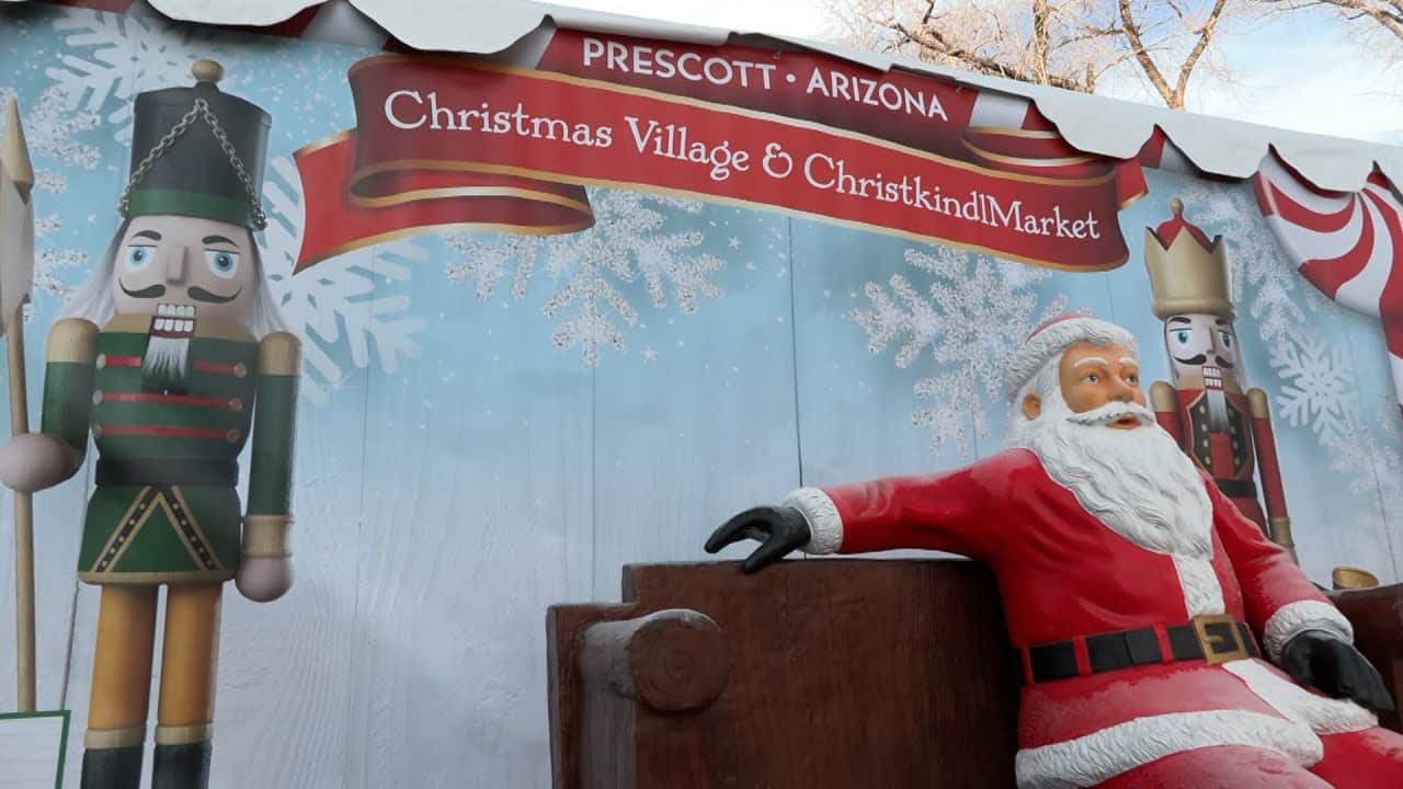 Christmas Markets Arizona 2025 Ultimate Guide to Festive Events, Lights & Holiday Shopping 1 A Santa Claus statue sits on a bench in front of a Christmas Village & Christkindl Market sign with nutcracker decorations in Prescott, Arizona.