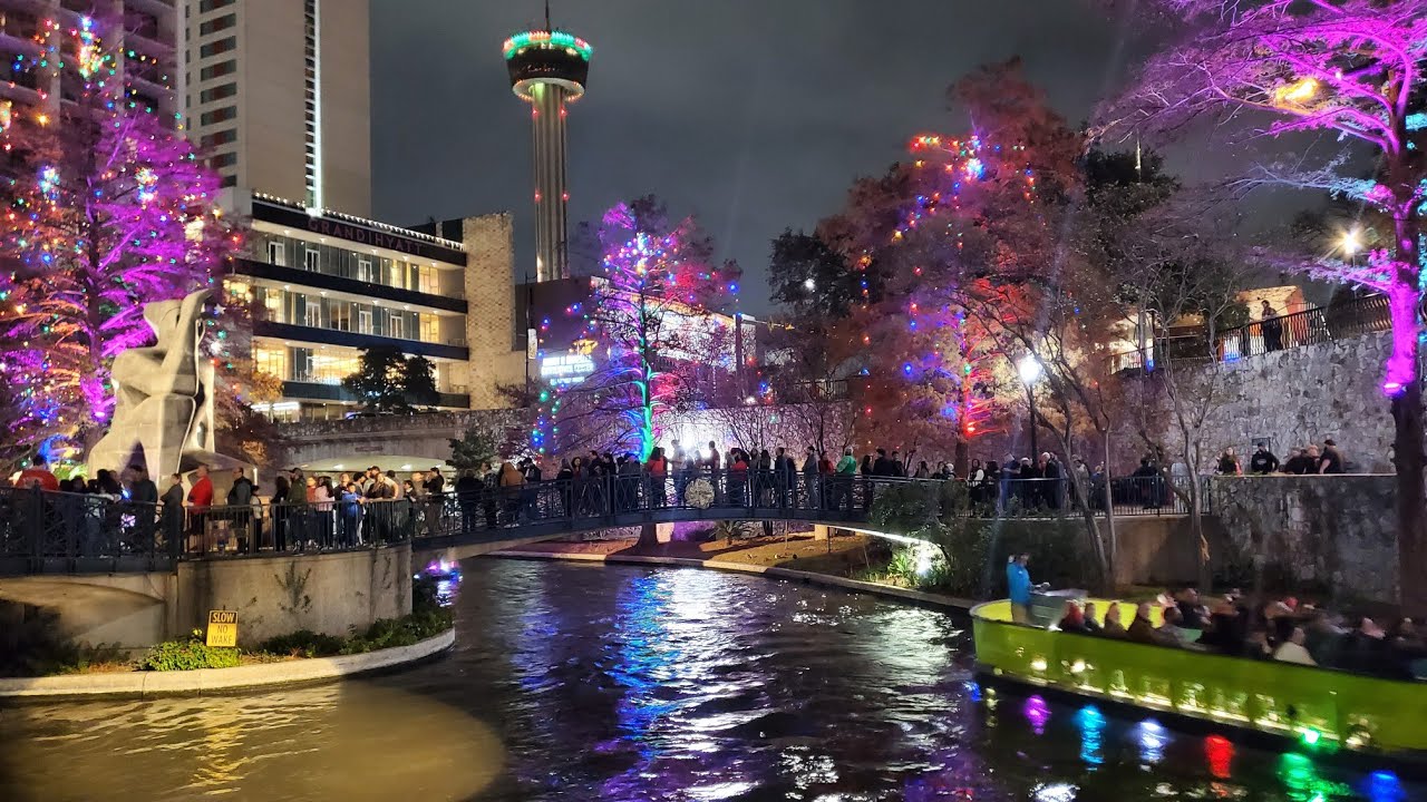 Christmas Markets Texas 2025: Top 50 Festive Destinations 1 A riverboat passes under a pedestrian bridge along a lively, illuminated riverside with crowds and colorful trees at night.
