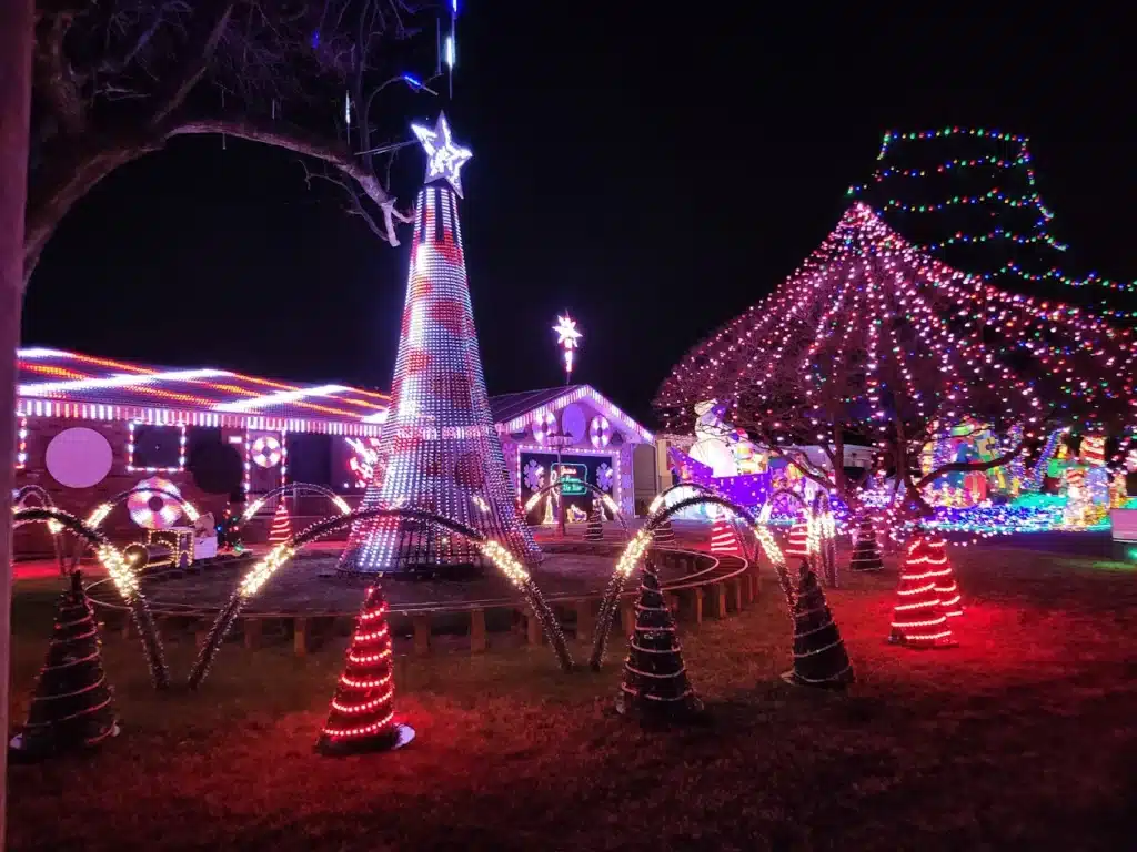 20 Magical Christmas Light Displays in Missouri You Can’t Miss This Holiday Season! (#6 is Jaw-Dropping) 1 A yard decorated for Christmas with colorful lights, including a tall cone-shaped tree with a star, illuminated arches, and lights covering a nearby tree and house.