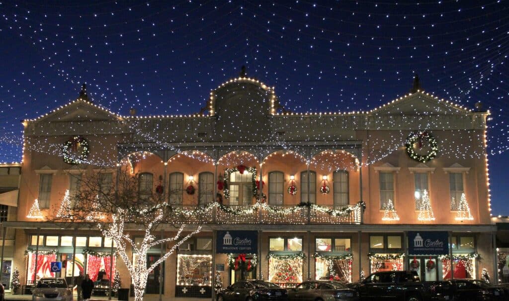 10 Jaw-Dropping Christmas Light Displays in Mississippi You Can’t Miss in 2025! 1 A two-story building decorated with Christmas lights and wreaths, with illuminated trees and shop windows, under a canopy of string lights at night.
