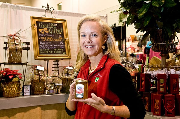 50 Magical Christmas Markets in Virginia for 2025 You Can’t Miss: Ultimate Guide to Festive Events & Holiday Shopping 6 A woman in a red vest holds a jar of jam while standing in front of a gift shop display with baskets, gift items, and a chalkboard sign.