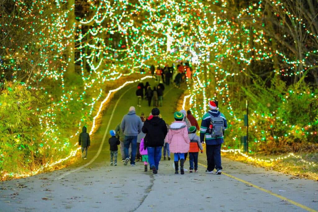 20 Magical Christmas Light Displays in Missouri You Can’t Miss This Holiday Season! (#6 is Jaw-Dropping) 4 A group of people walk along a paved path lined with trees and decorated with festive holiday lights at night.