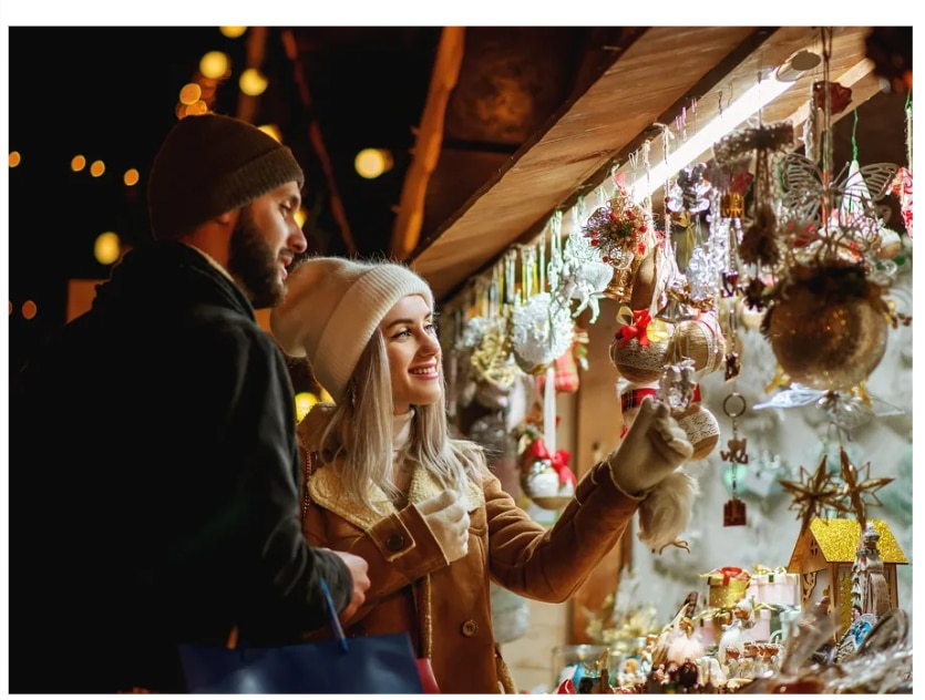 Christmas Markets Southern California 2025: Top 50 Festive Holiday Events and Locations 4 A man and a woman wearing winter clothing look at holiday ornaments at an outdoor market stall decorated with festive lights.