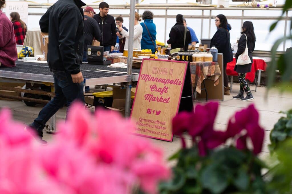 Minnesota’s 50 Best Christmas Markets to Visit in 2025 (You’ll Love #7!) 3 People browse vendor tables at an indoor market. A sign in the foreground reads "Welcome to Minneapolis Craft Market." Bright pink flowers are visible at the front.