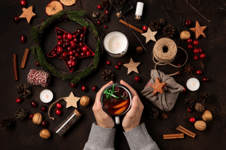 Christmas Markets Washington State 2025: Top 50 Festive Events and Holiday Fairs 2 Hands holding a mug of mulled wine surrounded by candles, cranberries, nuts, cinnamon sticks, cookies, and holiday decorations on a dark surface.