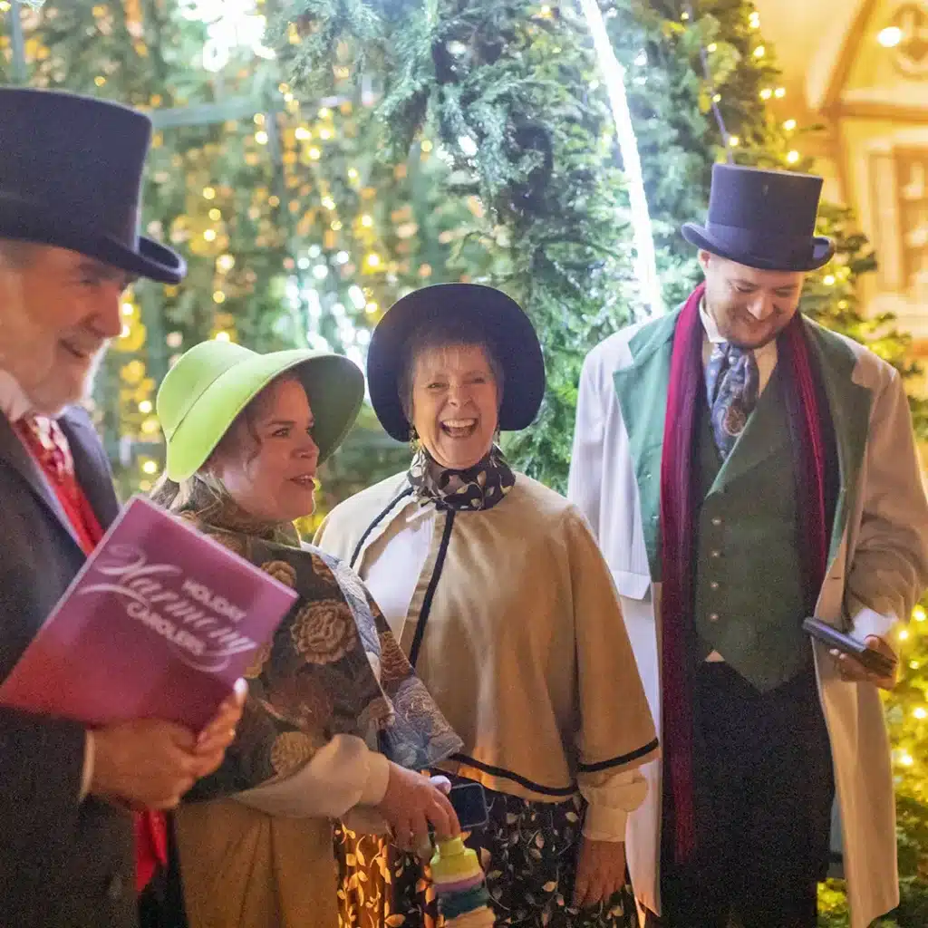 Christmas Markets Washington State 2025: Top 50 Festive Events and Holiday Fairs 6 Four adults in Victorian-style costumes stand together smiling under festive holiday lights and greenery, with one person holding a songbook.