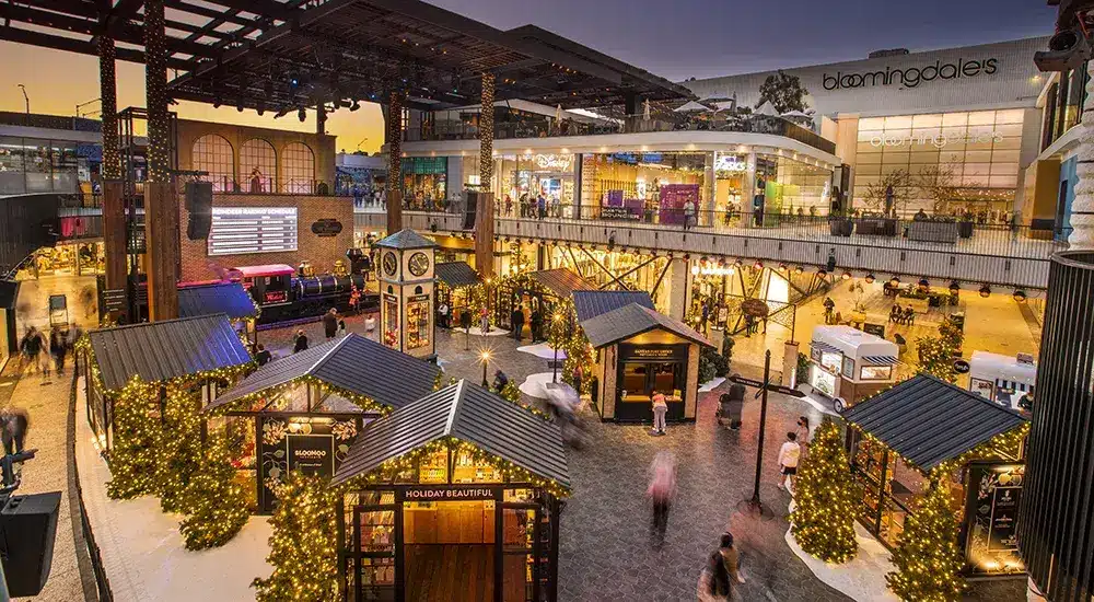 Christmas Markets Southern California 2025: Top 50 Festive Holiday Events and Locations 2 Outdoor shopping center decorated with holiday lights and small illuminated vendor huts, with people walking around and a Bloomingdale's store visible in the background.