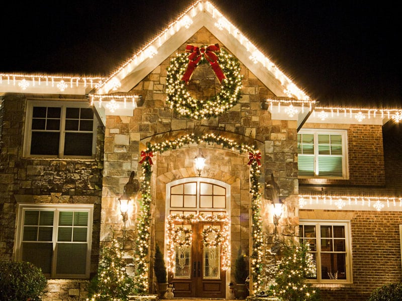 10 Dazzling Christmas Light Shows in Michigan You Can’t Miss in 2025 (Plus Free Spots & Secret Local Favorites!) 4 Two-story stone house decorated with strings of white Christmas lights and a large wreath with a red bow above the front door at night.
