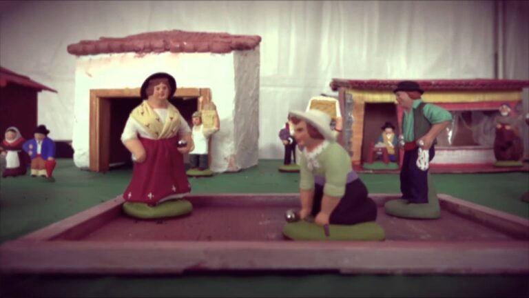 Clay figurines depict people playing a traditional outdoor game in a miniature village setting with houses and bystanders in the background.