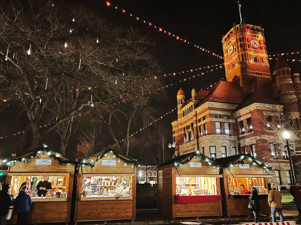 Ohio Christmas Markets 2025: 50 Unforgettable Holiday Events & Festive Fairs You Have to Visit 2 Four wooden holiday market stalls are lit up at night in front of a historic building with a clock tower, under strings of colorful lights and decorative icicles.