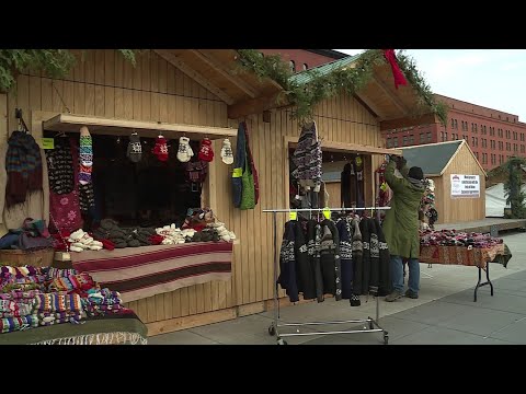 Outdoor market stalls with wooden huts display winter clothing, knitwear, and accessories; a person is arranging items on a rack.