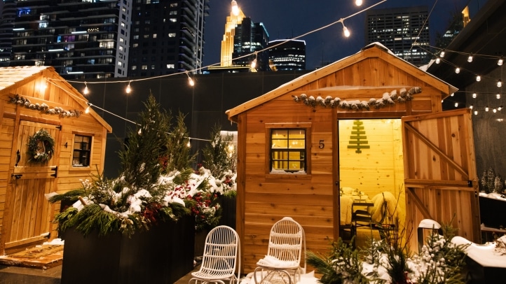 Minnesota’s 50 Best Christmas Markets to Visit in 2025 (You’ll Love #7!) 4 Two small wooden cabins decorated with wreaths and string lights sit among snowy plants, with city skyscrapers visible in the background at night.