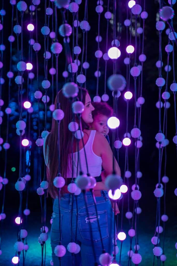 10 Dazzling Christmas Light Shows in Michigan You Can’t Miss in 2025 (Plus Free Spots & Secret Local Favorites!) 5 A woman holding a child stands among hanging strands of glowing purple lights at night.