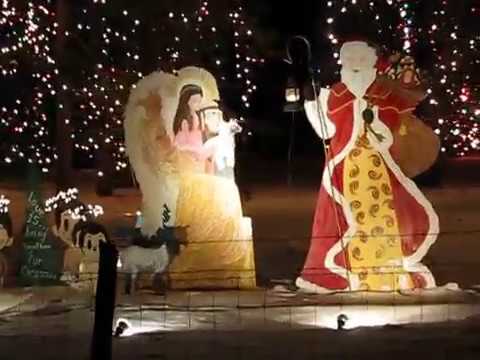 A holiday display features painted cutouts of Santa Claus approaching a nativity scene with Mary, baby Jesus, and an angel, with Christmas lights in the background.