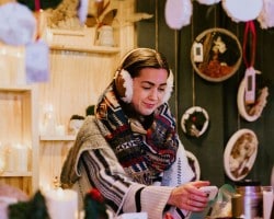Minnesota’s 50 Best Christmas Markets to Visit in 2025 (You’ll Love #7!) 3 A woman wearing earmuffs and a patterned scarf stands indoors, smiling while using a smartphone. Shelves with various decorative items are visible in the background.