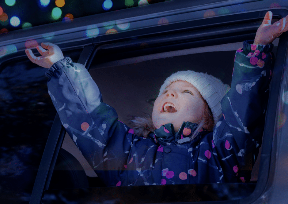 7 Jaw-Dropping Christmas Light Shows in Nebraska You Can’t Miss This Holiday Season (2025 Guide!) 4 A child wearing a winter coat and hat leans out of a car window, smiling with arms raised, with colorful lights visible in the background.