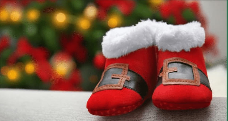A pair of red baby booties with white fur trim and black belts, designed to resemble Santa’s boots, is shown in front of a blurred festive background.
