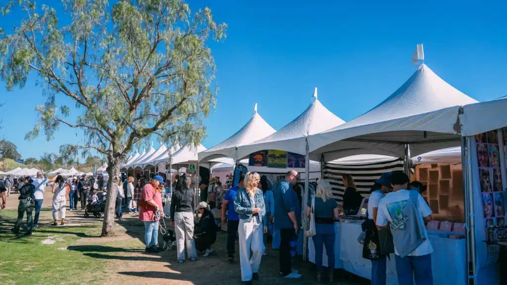 Christmas Markets Southern California 2025: Top 50 Festive Holiday Events and Locations 3 People visit vendor tents at an outdoor market or festival under clear blue skies, with a tree and grassy area visible on the left.