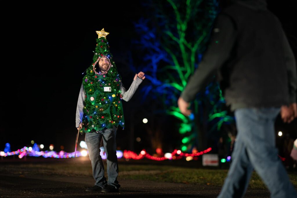10 Dazzling Christmas Light Shows in Michigan You Can’t Miss in 2025 (Plus Free Spots & Secret Local Favorites!) 6 A person dressed as a decorated Christmas tree stands outdoors at night, with festive lights and two people walking nearby.