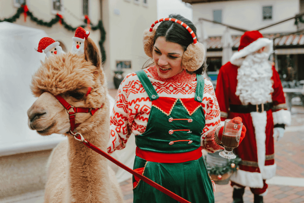 Minnesota’s 50 Best Christmas Markets to Visit in 2025 (You’ll Love #7!) 3 A woman in festive holiday attire holds a drink and leads a llama with antler decorations; a person dressed as Santa Claus stands in the background.