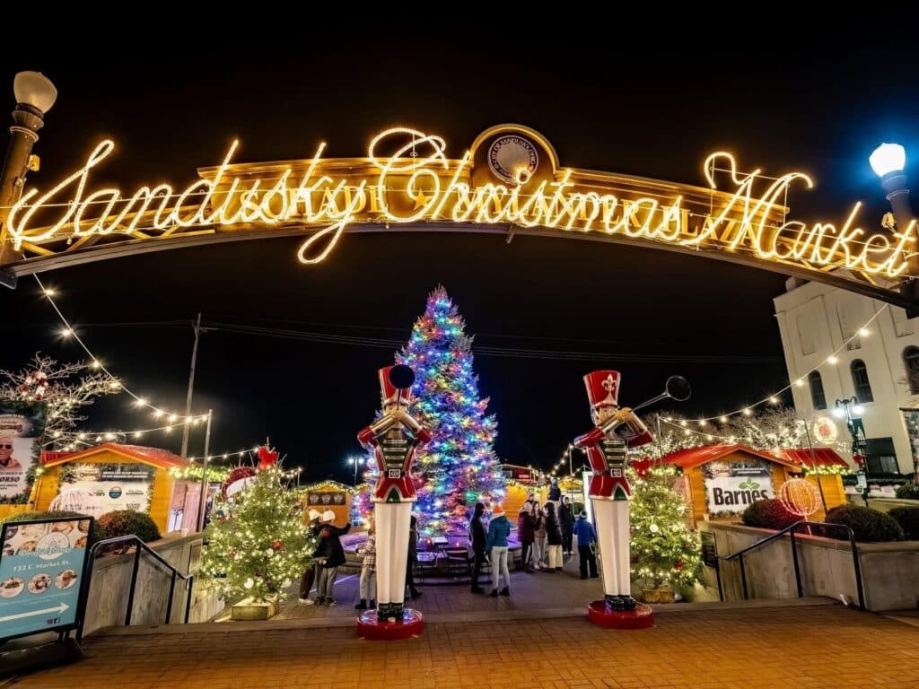 Ohio Christmas Markets 2025: 50 Unforgettable Holiday Events & Festive Fairs You Have to Visit 3 Illuminated sign reading "Sandusky Christmas Market" at night with a large decorated Christmas tree, toy soldier statues, market stalls, and people in the background.