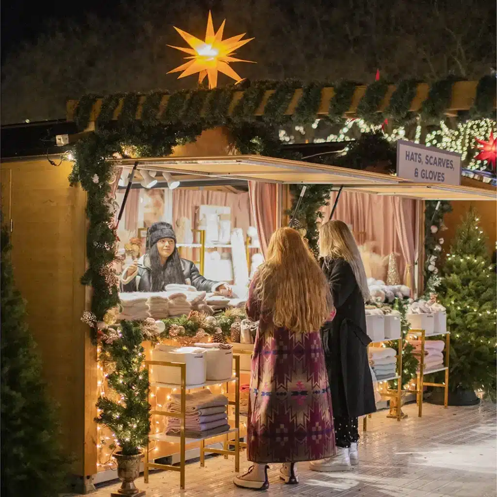 Christmas Markets Washington State 2025: Top 50 Festive Events and Holiday Fairs 5 Two women shop for winter accessories at an outdoor market stall decorated with greenery and lights. The vendor stands behind a counter displaying hats, scarves, and gloves.
