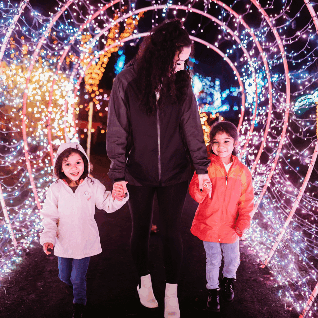 Christmas Markets Washington State 2025: Top 50 Festive Events and Holiday Fairs 3 An adult and two children walk hand in hand through a tunnel of colorful, glowing holiday lights at night.