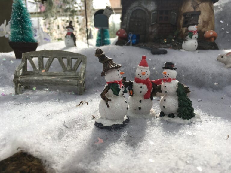 Three small snowman figurines stand together on artificial snow near a wooden bench, with miniature trees and a house in the background.