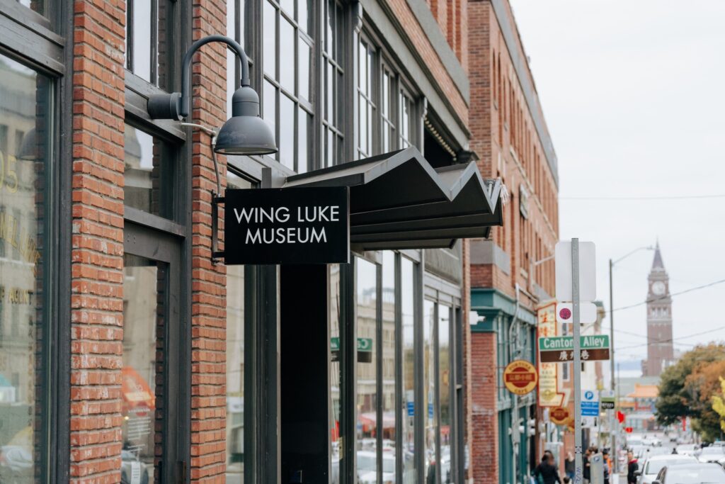 Christmas Markets Washington State 2025: Top 50 Festive Events and Holiday Fairs 8 A black sign reading "Wing Luke Museum" hangs from a brick building along a city street with other storefronts and a distant clock tower visible.