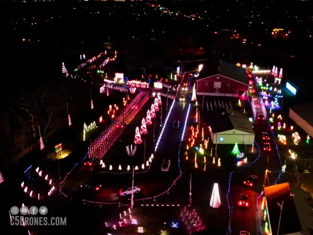 7 Jaw-Dropping Christmas Light Displays in New Jersey That Will Blow Your Mind (And Melt Your Frosty Heart!) 4 Aerial view of a drive-through holiday light display at night, with decorated trees, illuminated figures, and cars moving along marked paths.