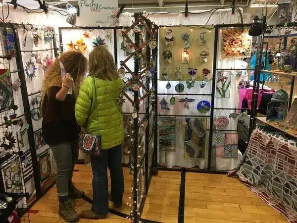 50 Magical Christmas Markets in Virginia for 2025 You Can’t Miss: Ultimate Guide to Festive Events & Holiday Shopping 6 Two people stand in front of a booth displaying various colorful stained glass art pieces at an indoor craft fair.