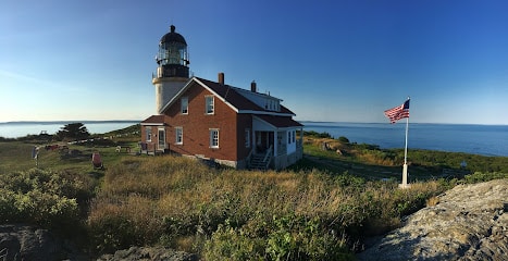 Maine’s Ultimate 2025 Christmas Light Guide: Discover Hidden Sparkling Gems & Holiday Magic Displays You Can’t Miss! 4 A brick lighthouse and house sit on a grassy coastal bluff with an American flag in the foreground and the ocean in the background under a clear blue sky.