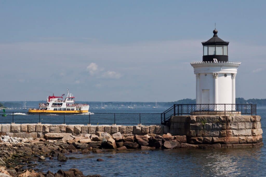 Maine’s Ultimate 2025 Christmas Light Guide: Discover Hidden Sparkling Gems & Holiday Magic Displays You Can’t Miss! 1 A white lighthouse stands on a rocky shoreline in the foreground, while a ferry boat travels across the water in the background under a clear sky.