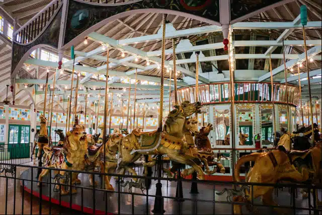 Louisiana’s Ultimate 2025 Christmas Light Displays: Discover The Most Magical, Must-See Holiday Shows You Can’t Miss! 10 An indoor carousel with ornate, painted horses and mirrors, enclosed by a fence, under a high, wooden ceiling with exposed beams.