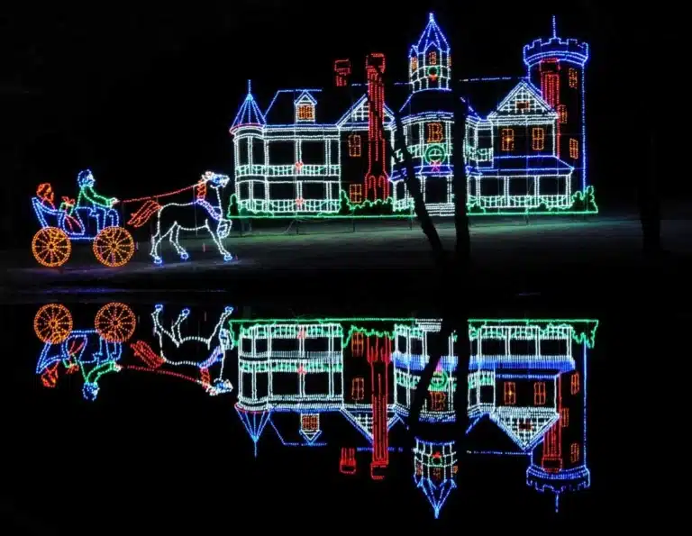 Holiday lights display of a horse-drawn carriage passing a large castle, with colorful lights reflected in water below at night.