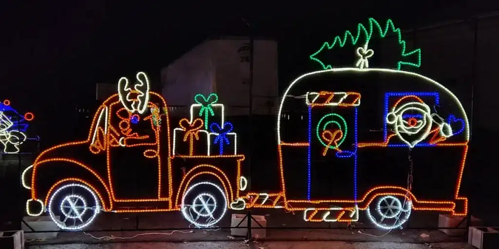 18 Magical Christmas Light Displays in Wisconsin You Can’t Miss in 2025 (Drive-Thrus, Walk-Thrus & Secret Neighborhood Gems!) 5 A holiday light display of a truck with a reindeer driving, towing a trailer with Santa inside and a Christmas tree on top, both decorated with presents and festive designs.