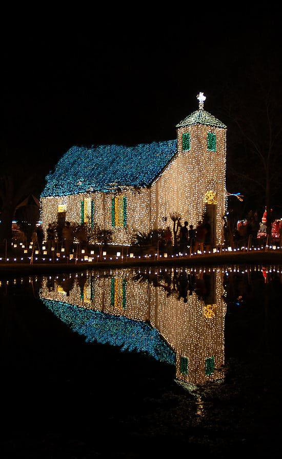 A church decorated with blue and white Christmas lights is reflected in a calm body of water at night.