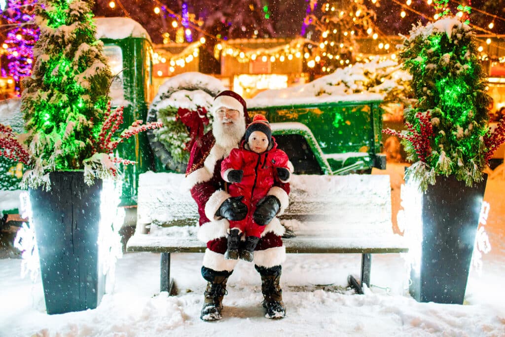18 Magical Christmas Light Displays in Wisconsin You Can’t Miss in 2025 (Drive-Thrus, Walk-Thrus & Secret Neighborhood Gems!) 2 Santa Claus sits on a snowy bench with a young child in a red coat on his lap, surrounded by festive lights and decorated planters outdoors at night.