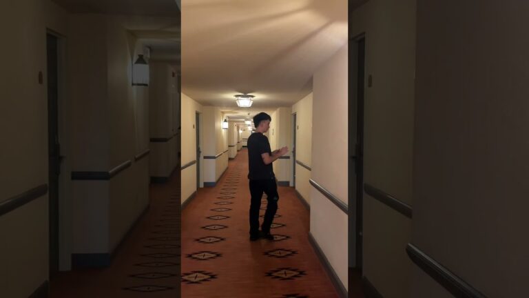A person dressed in black stands in the hallway of a building, facing sideways while looking at a phone or tablet. The corridor is lit with ceiling lamps and lined with doors.