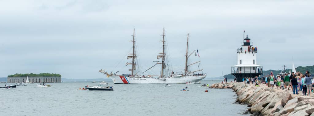 Maine’s Ultimate 2025 Christmas Light Guide: Discover Hidden Sparkling Gems & Holiday Magic Displays You Can’t Miss! 2 A large white Coast Guard tall ship sails near a lighthouse as people gather on a rocky pier and small boats float on the water under a cloudy sky.