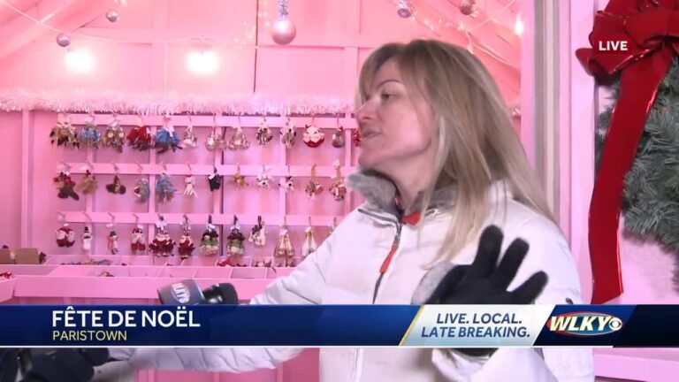 A woman in a white jacket speaks during a news interview at a decorated pink holiday market booth with ornaments hanging on the wall.