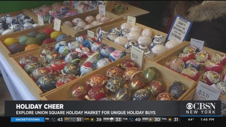 Painted decorative ornaments are displayed in wooden trays at a market booth, with price tags and a sign requesting customers not to handle items without staff assistance.
