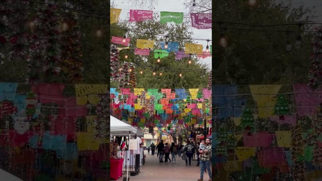 Colorful papel picado banners hang above an outdoor market with people walking among stalls and trees in the background. String lights are also visible overhead.